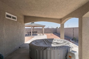 A covered outdoor area with a hot tub, a patio with chairs, and a view of a building in the background.