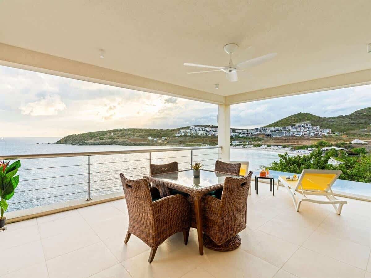 Ein Balkon mit Tisch und Stühlen, einem Deckenventilator und Blick auf das Meer und die Hügel.