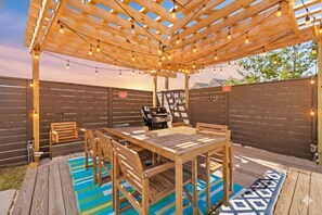 Un patio exterior con una pérgola de madera, luces colgantes, una parrilla y una mesa de comedor con sillas.