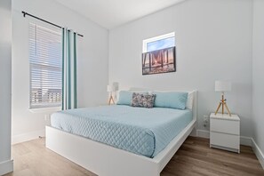 Un dormitorio con una cama, dos mesitas de noche, una ventana con persianas y un cuadro en la pared.
