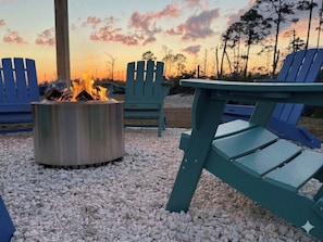 A fire pit surrounded by Adirondack chairs on a gravel surface with a sunset in the background.