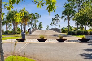 A paved pathway leading to a bridge with a view of a park with various trees and planters. - Le Bleu Bebe 5BR | Golf Cart + Game Room + Bikes (Port St. Joe)