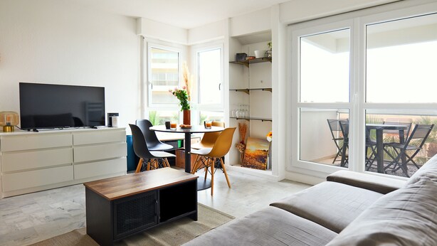 Una sala de estar moderna con un televisor de pantalla plana, un comedor con mesa y sillas, y un ventanal con vistas al exterior.