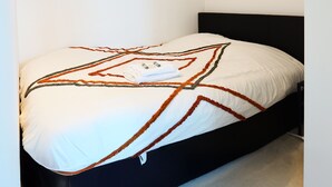 Una cama bien hecha con una colcha blanca y marrón de estampado geométrico, una toalla doblada y dos botellas de loción sobre la cama.