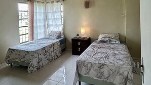 A bedroom with two beds, wooden ceiling beams, a small bedside table with a lamp, and a window with curtains.