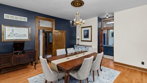 A dining room with a wooden table, white chairs, a chandelier, and framed pictures on the walls.