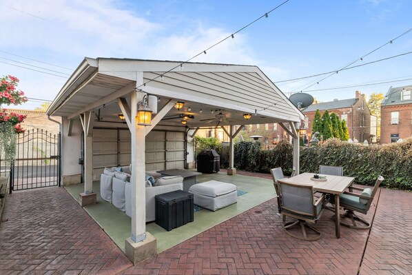 Een terras buiten met een overdekte zithoek, een tafel met stoelen en een barbecue.