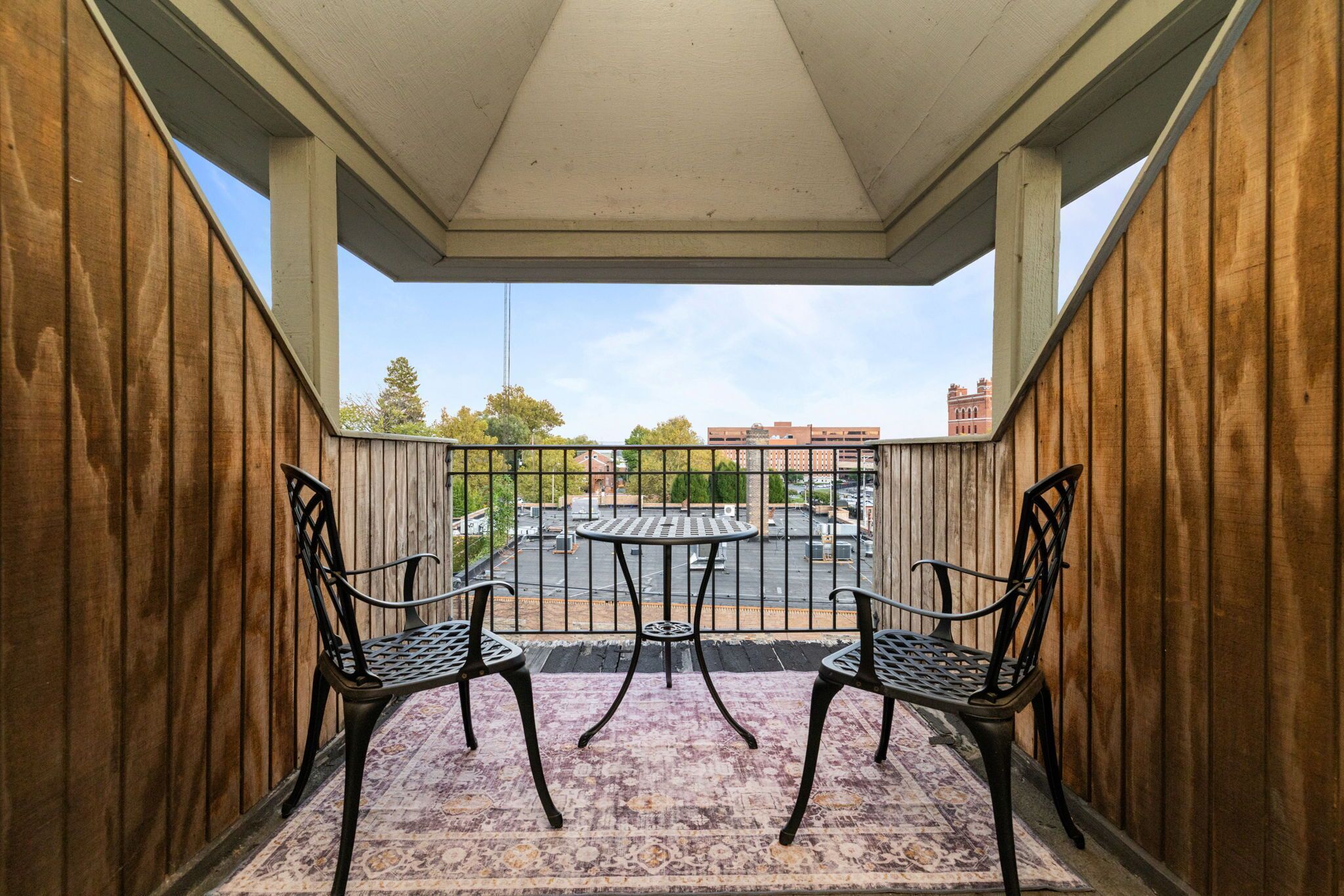 A covered balcony with two metal chairs and a small table, overlooking a parking area and buildings.