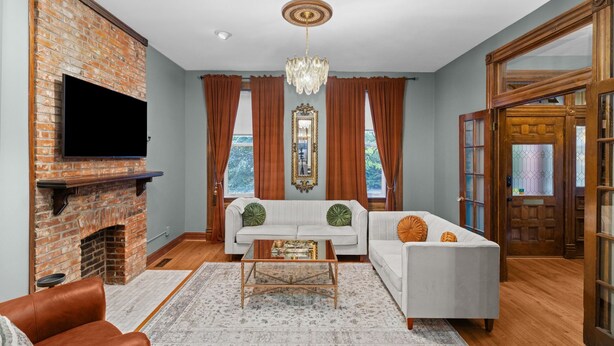 A living room with a brick fireplace, white sofas, a chandelier, and a large window with curtains.