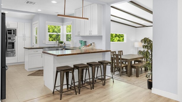 A modern kitchen with white cabinetry, a wooden island with bar stools, a dining area with a wooden table and chairs, and a view of greenery outside. - UPSCALE & UPGRADED 3BR HOUSE in ENCINO with GARAGE & LARGE FENCED YARD/PATIO p84 (Los Angeles)