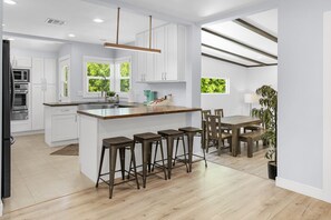 A modern kitchen with white cabinetry, a wooden island with bar stools, a dining area with a wooden table and chairs, and a view of greenery outside. - UPSCALE & UPGRADED 3BR HOUSE in ENCINO with GARAGE & LARGE FENCED YARD/PATIO p84 (Los Angeles)