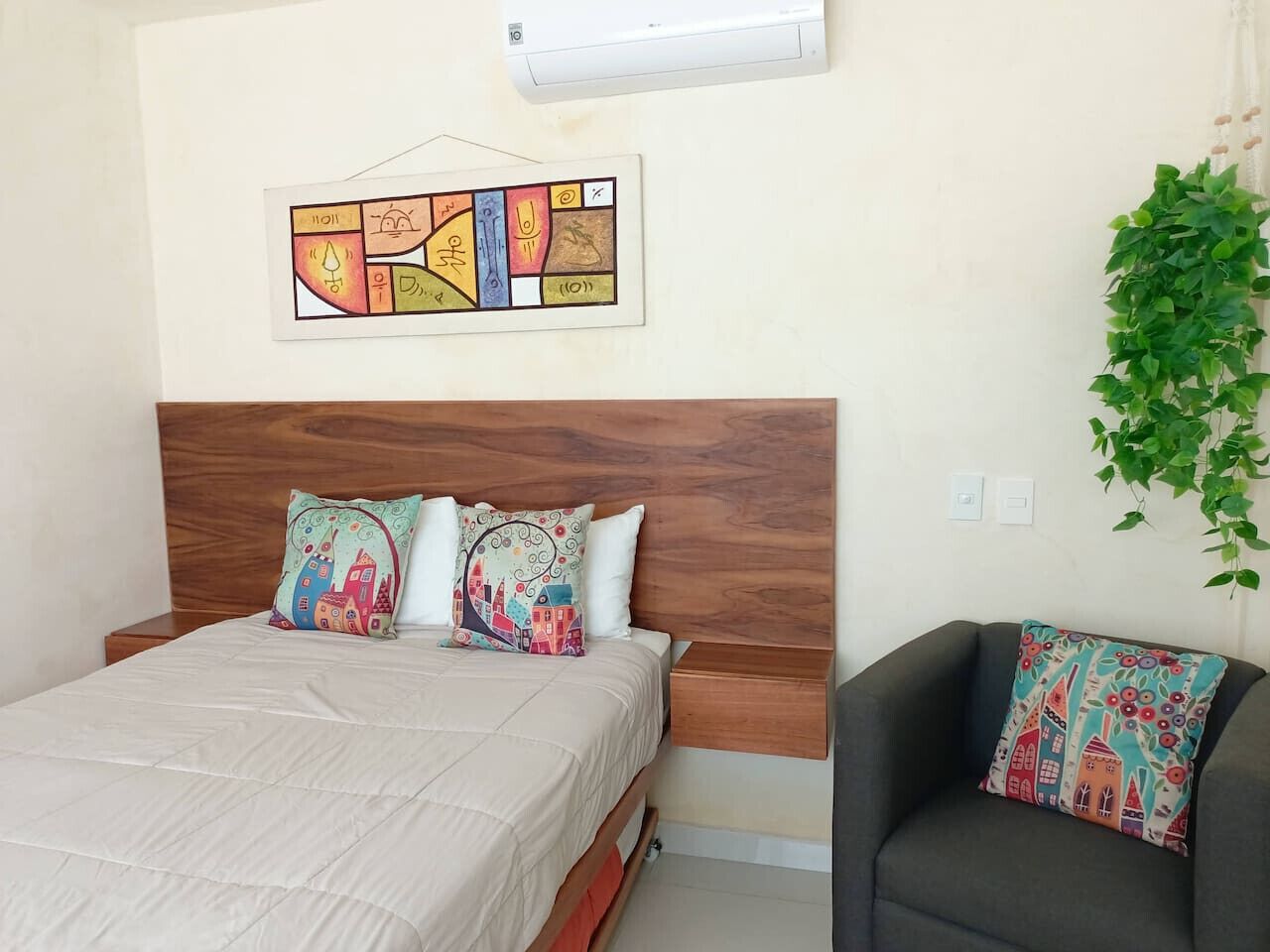 Une chambre à coucher avec une tête de lit en bois, un lit agrémenté d’oreillers à motifs, un fauteuil avec un coussin et un climatiseur fixé au mur.