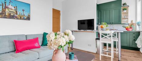 A modern living room with a grey sofa, a pink pillow, a vase with flowers, and a flat-screen TV.