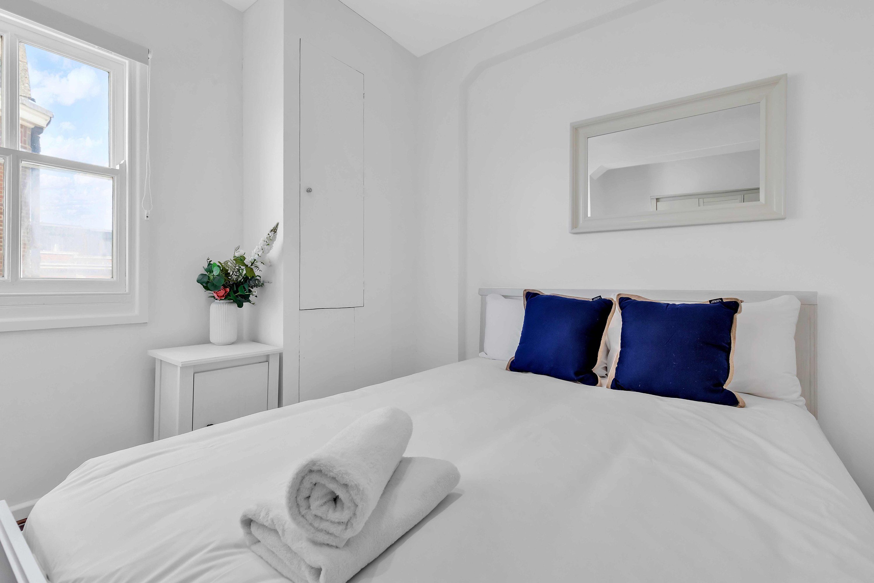 Una cama bien tendida con ropa de cama blanca y toallas enrolladas, una mesita de noche con una planta y una ventana con vista al cielo.