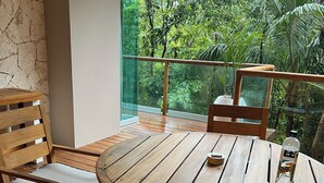 Una terraza de madera con una mesa redonda y sillas, una botella de agua y vistas a exuberante follaje a través de una barandilla de cristal.