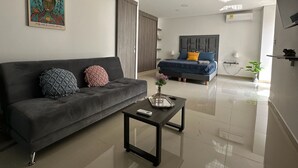 A modern living room with a grey sofa, a small coffee table, and a bed with blue and yellow pillows. - Nirvana ApartaHotel (Cali)