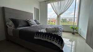 A bedroom with a bed, a window with curtains, a potted plant, and a view of buildings. - Nirvana ApartaHotel (Cali)