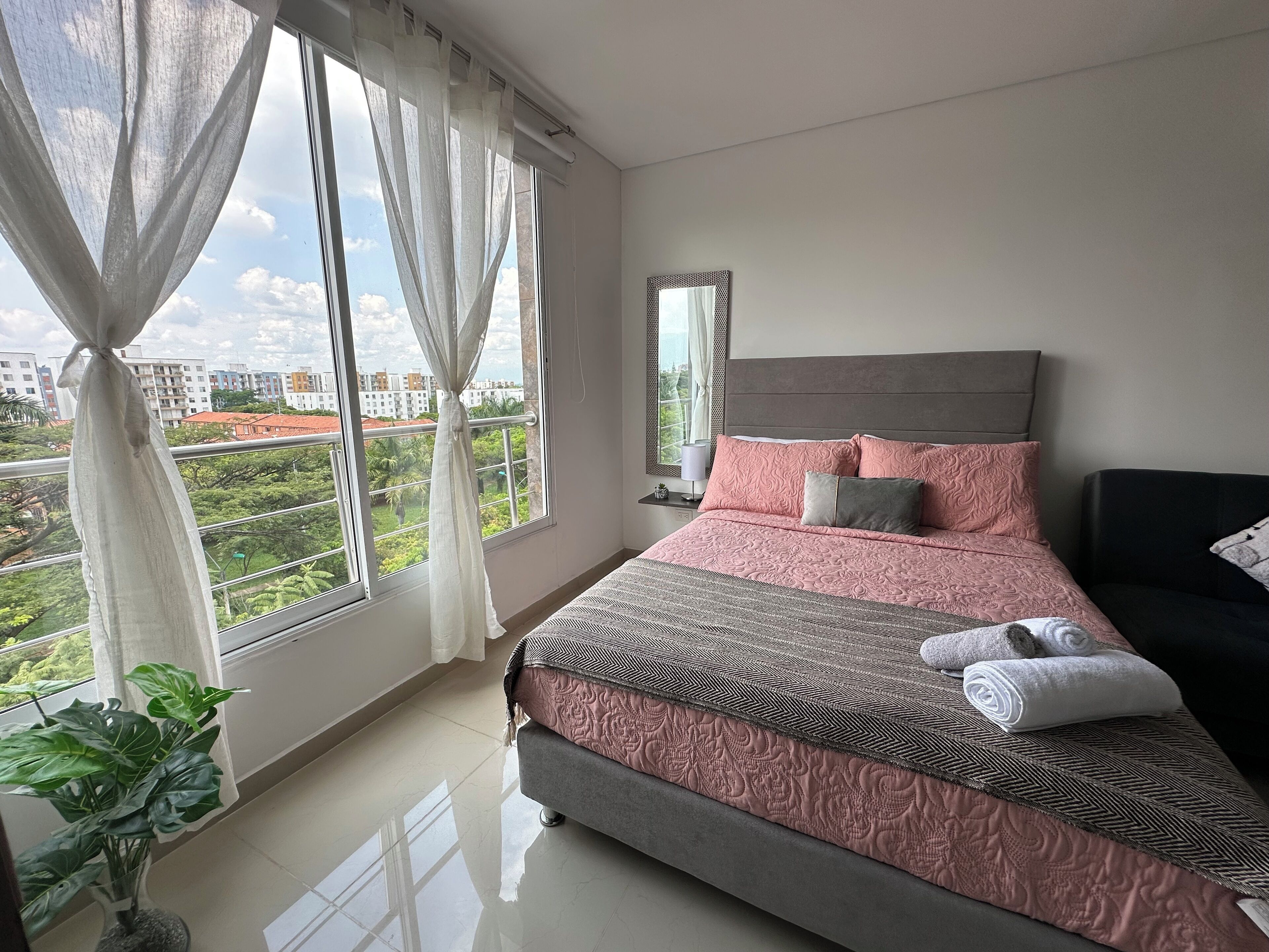 A bedroom with a large bed, a bedside table, a mirror, and a view of greenery outside.