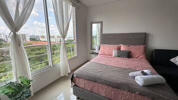 A bedroom with a large bed, a bedside table, a mirror, and a view of greenery outside.