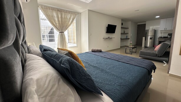 A modern bedroom with a large bed, a TV mounted on the wall, and a kitchenette in the background. - Nirvana ApartaHotel (Cali)