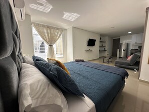Un dormitorio moderno con una cama grande, un televisor montado en la pared y una kitchenette al fondo.