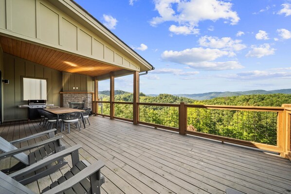 A deck with a wooden table and chairs, a grill, and a view of a forested mountain range.