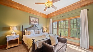 A bedroom with a large bed, a wooden ceiling fan, a nightstand with a lamp, a window with curtains, and a view of trees. - Mountain Home with Mountain Views (Sugar Mountain)
