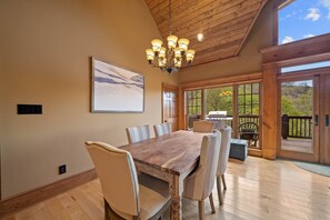 A dining area with a wooden table and chairs, a chandelier, and a large window with a view of greenery.