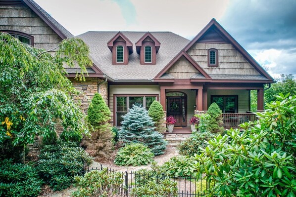 A two-story house with a stone facade, a front porch, and a well-maintained garden with various shrubs and a small tree.
