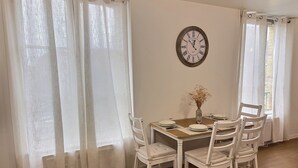 A dining area with a wooden table, white chairs, a vase with flowers, and a wall clock. - Entre Port & Mer - Spacious 77m2 apartment in Dieppe (Dieppe)