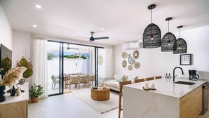 Una cucina moderna con isola centrale, una zona pranzo con porta in vetro che si apre su un balcone e un soggiorno con divano e tavolino.
