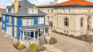 A two-story building with a blue exterior and a red roof, featuring a porch with outdoor seating and a small garden area. - Elegant Decor, Plush Bedding! Maya Angelou Room (Olean)