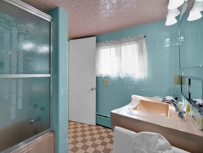 Une salle de bain aux murs de couleur turquoise, avec une porte blanche, un carrelage à motifs, un lavabo avec un miroir et une fenêtre ornée d’un rideau blanc.