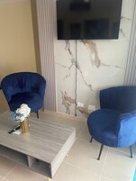 Un salón moderno con dos sillones azules, una mesa de centro con un jarrón de flores y un televisor de pantalla plana montado en una pared de mármol.