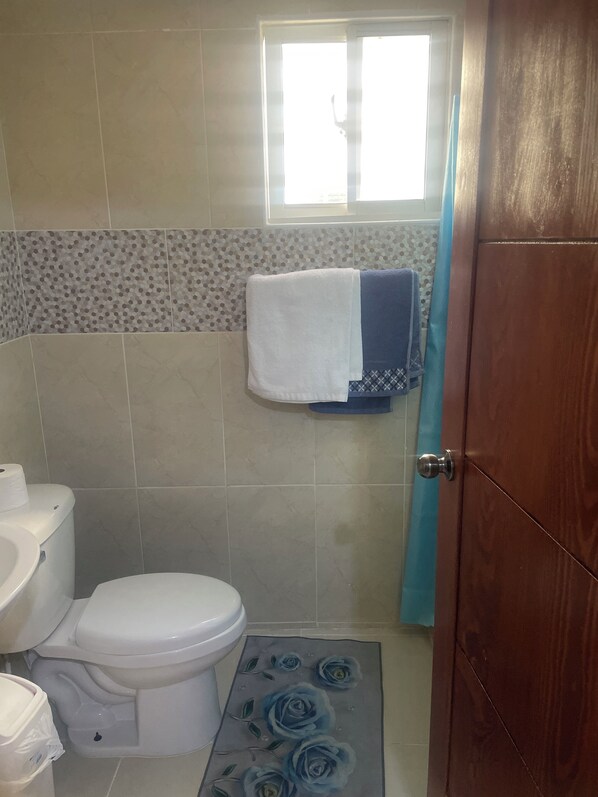 A bathroom with a toilet, a sink, a shower curtain, and a towel rack with towels. - POSADA DEL CARIBE (Santo Domingo Este)