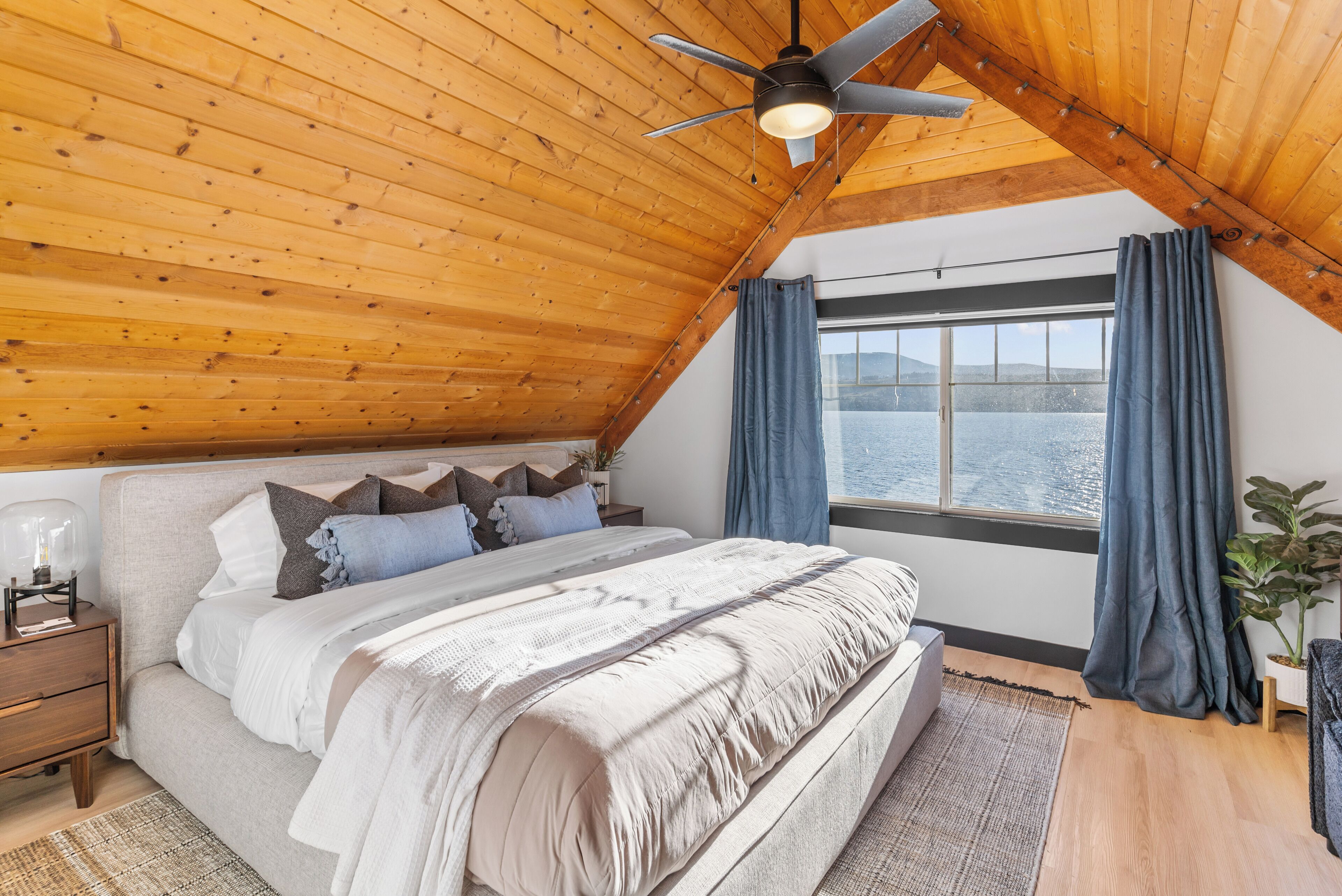 A bedroom with a wooden ceiling, a bed with a white comforter, a nightstand, a window with curtains, and a view of the water outside.