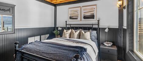 Um quarto com teto de madeira, uma cama com cabeceira escura, um criado-mudo com abajur e uma janela com vista.