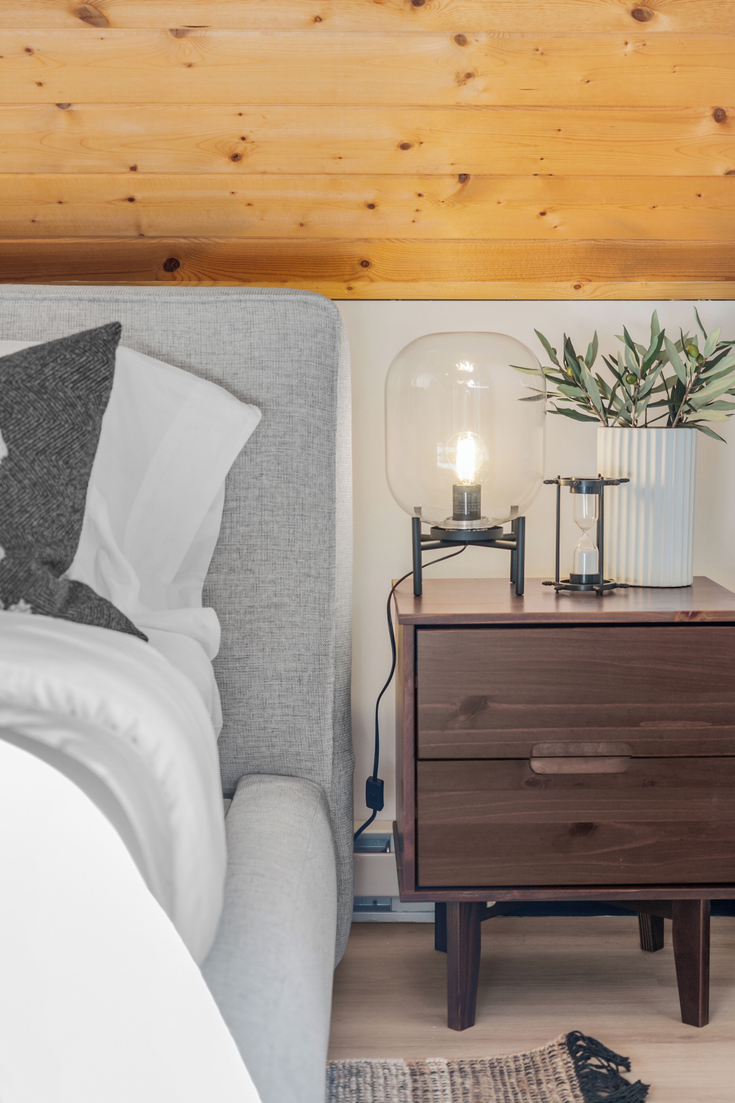 Un dormitorio con techo de madera, un sofá gris, una mesita de noche con lámpara y una planta en maceta.