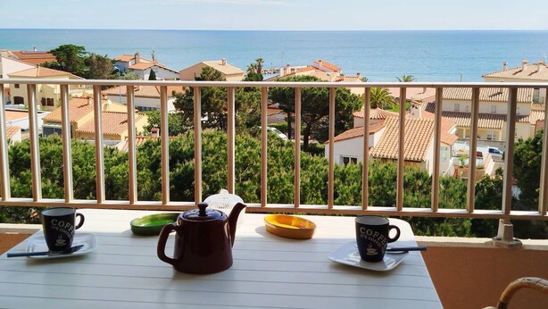 A balcony with a table set for coffee, overlooking a coastal town with terracotta-roofed buildings.