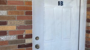Une porte blanche portant le numéro 9B, une poignée de porte en métal et un mur de briques.