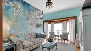 A living room with a beige sofa, a teal coffee table, and a large floral painting on the wall. - Signature Suite, 1895 Victorian Old Library Inn! (Olean)