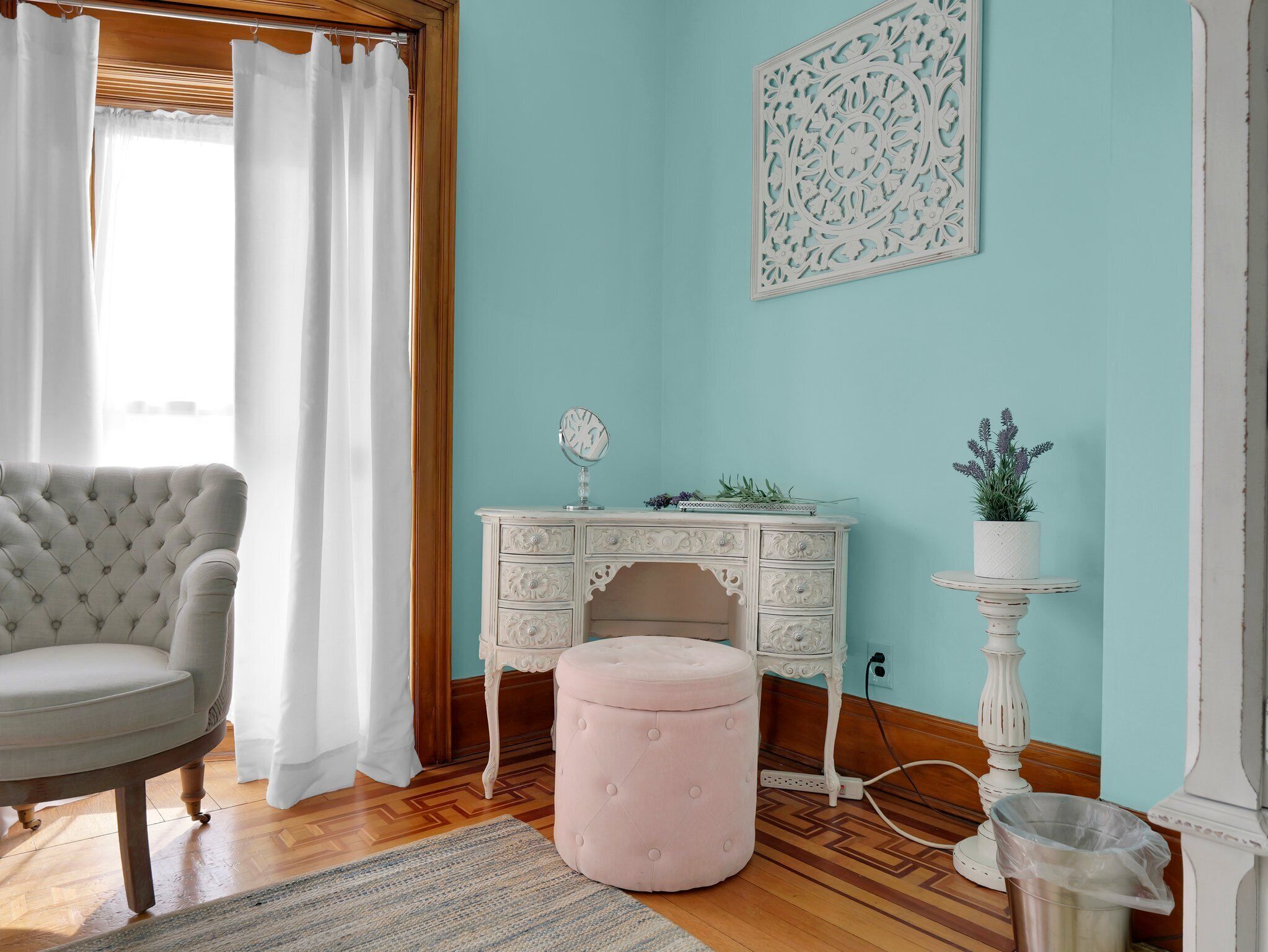 A room with a tufted armchair, a white dresser with floral carvings, a pink ottoman, a small white table with a plant, and a decorative wall piece.