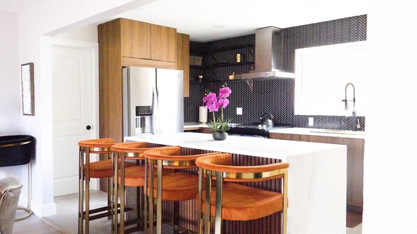 A modern kitchen with a central island, bar stools, and a decorative plant.