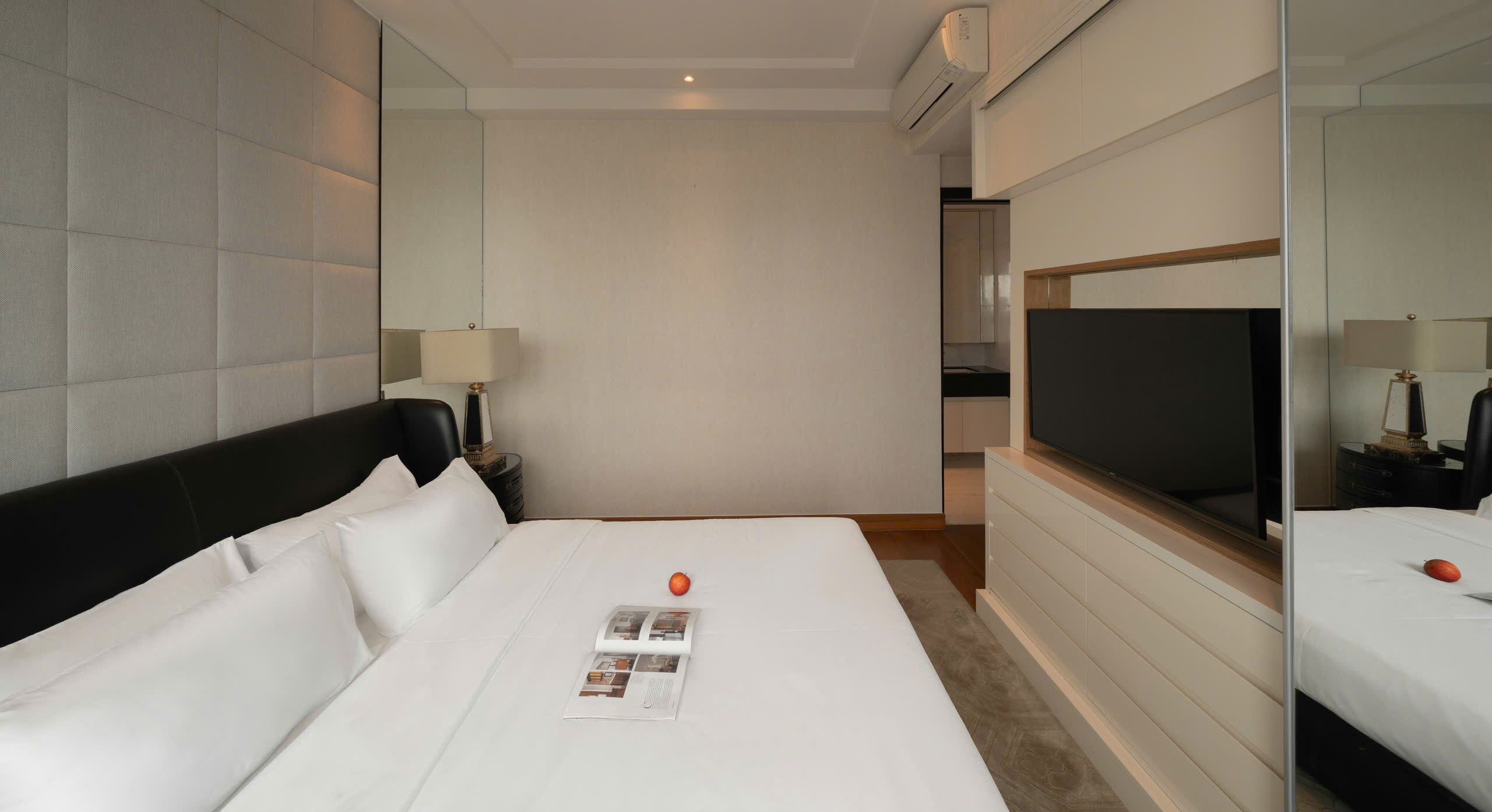 A modern hotel room with a large bed, a flat-screen TV, a bedside table with a lamp, and a small kitchen area in the background.