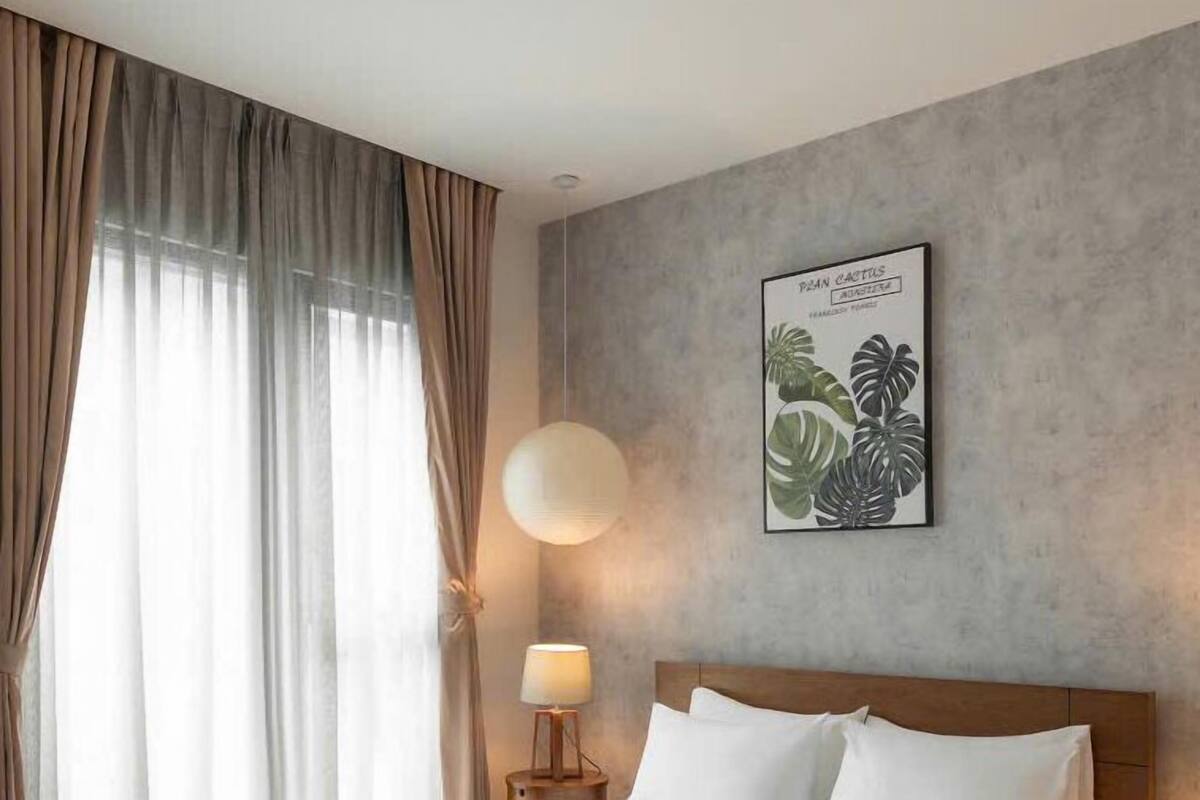 Un cuarto de hotel con una cama grande, una mesita de noche con lámpara, un grabado botánico enmarcado en la pared y cortinas translúcidas en la ventana.