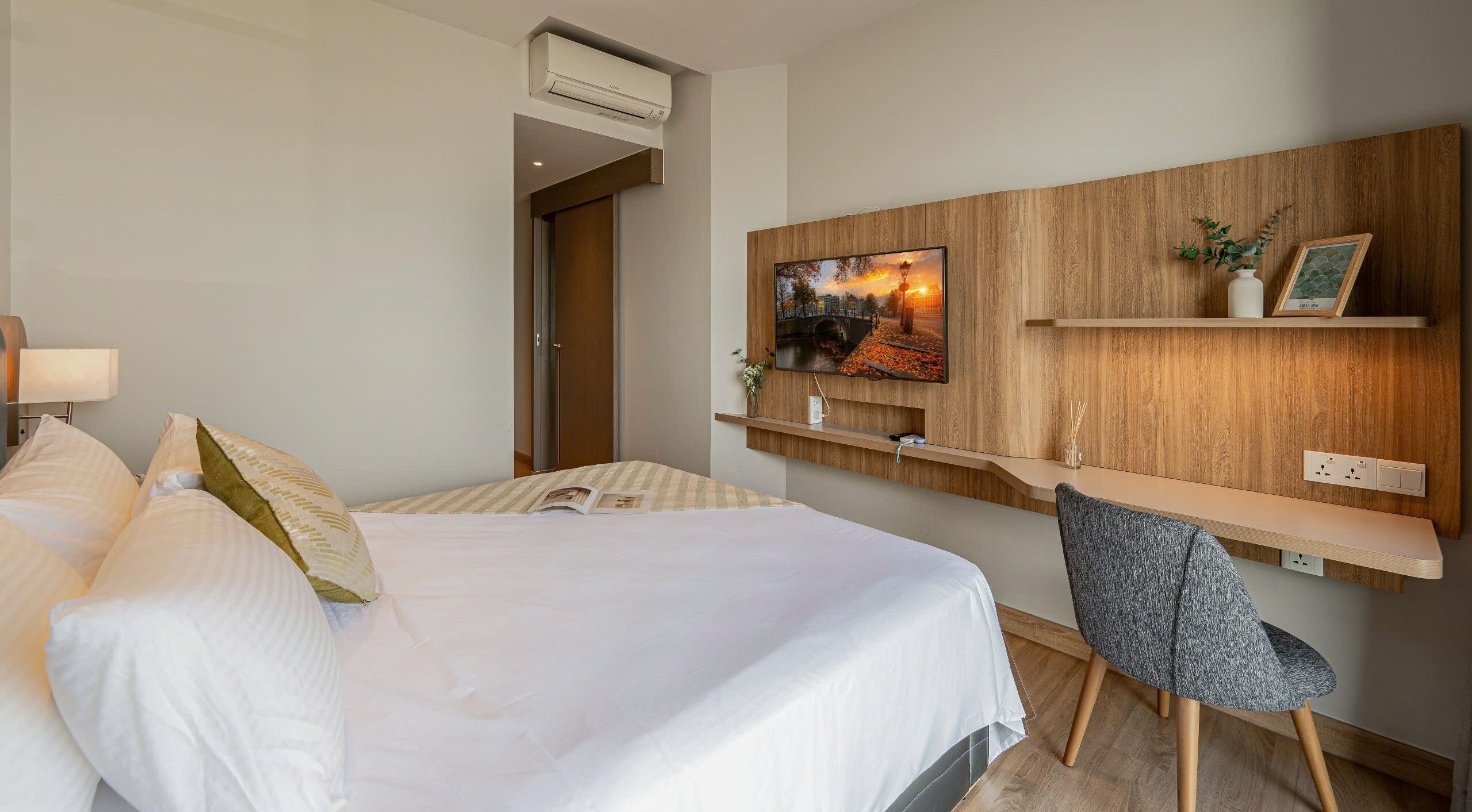 Ein Hotelzimmer mit einem Bett, einem Schreibtisch, einem Stuhl, einem an einer Holzvertäfelung montierten Fernseher und einer Tür zu einem weiteren Raum.