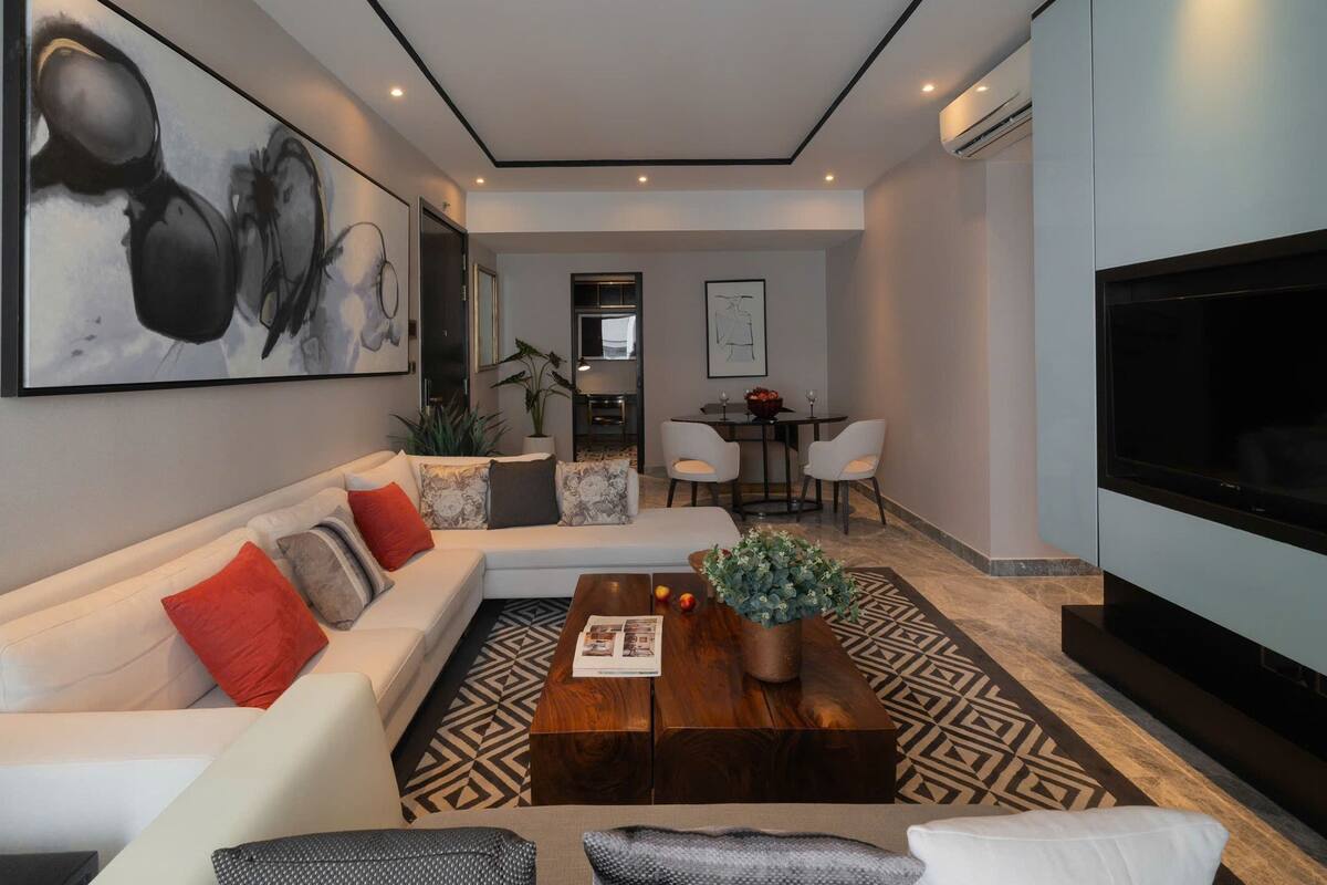 Una sala de estar moderna con un sofá, una mesa de centro y un televisor grande. En el fondo, hay un área de comedor con sillas y una mesa.
