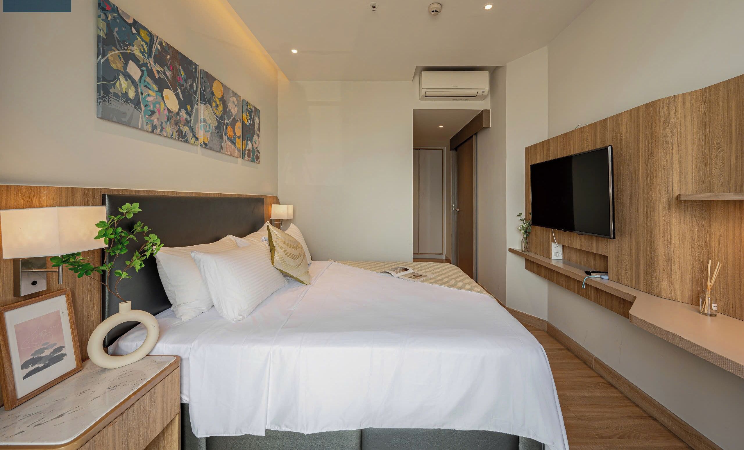 Een moderne hotelkamer met een groot bed, een flatscreen televisie aan de muur, een nachtkastje met een lamp, en een groot abstract schilderij boven het bed.