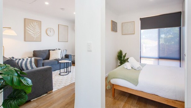 A modern, well-lit room with a grey sofa, a small round table, and a bed with a green blanket. There are framed artworks on the walls and a large window providing natural light. - Little Fig Retreat - 5 mins to Fremantle (Samson)