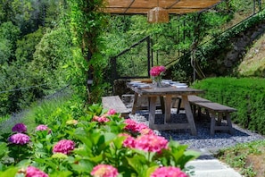 A wooden outdoor dining area with a table and benches, surrounded by lush greenery and flowers.